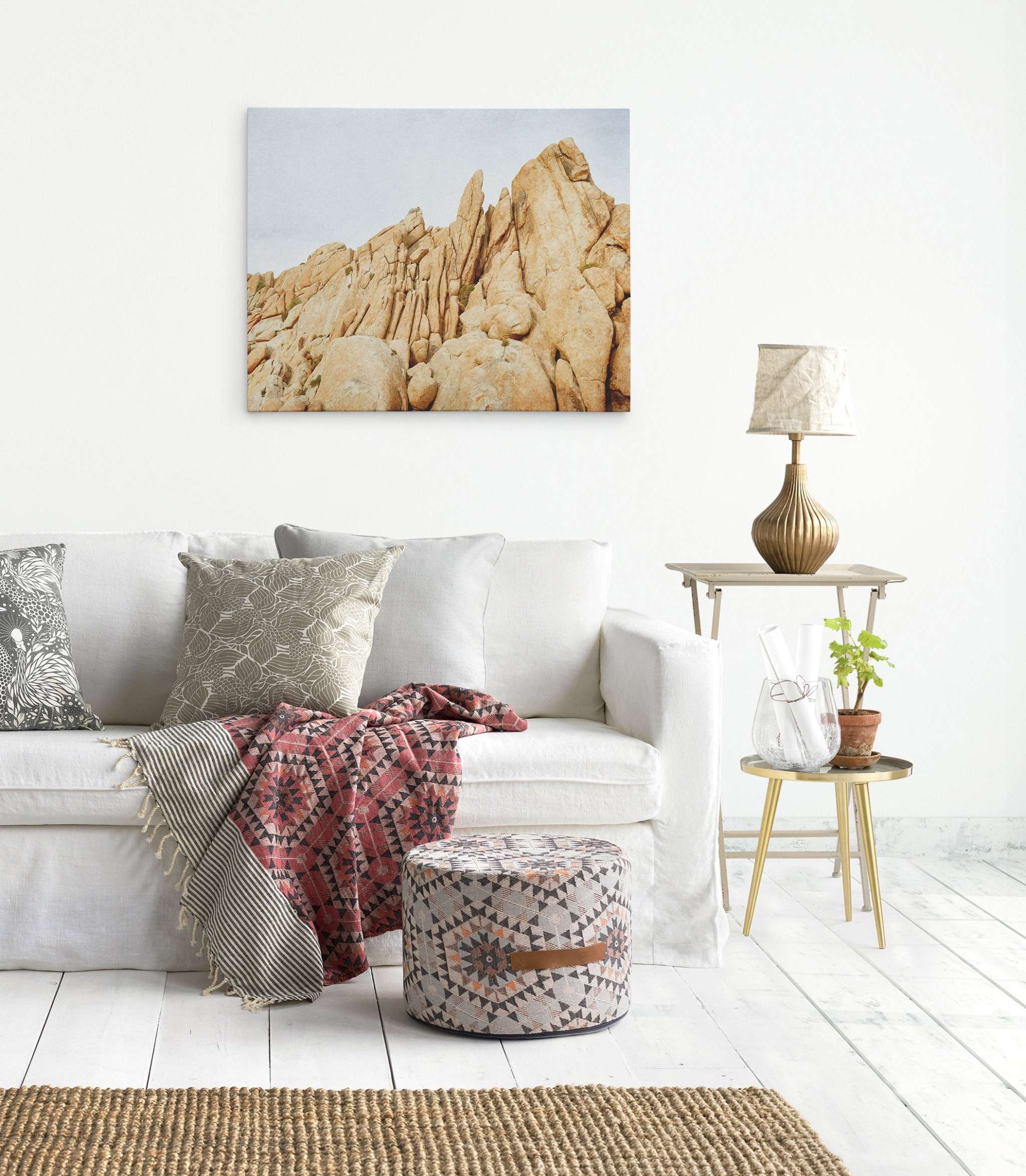 A modern living room with a white sofa adorned with various patterned cushions and a red geometric throw. A round patterned pouf is in front of the sofa. The wall showcases Offley Green's Joshua Tree Canvas Wall Art, 'Joshua Rocks,' depicting yellow desert rock formations. A small side table with a lamp, plant, and glass bottle completes the scene.