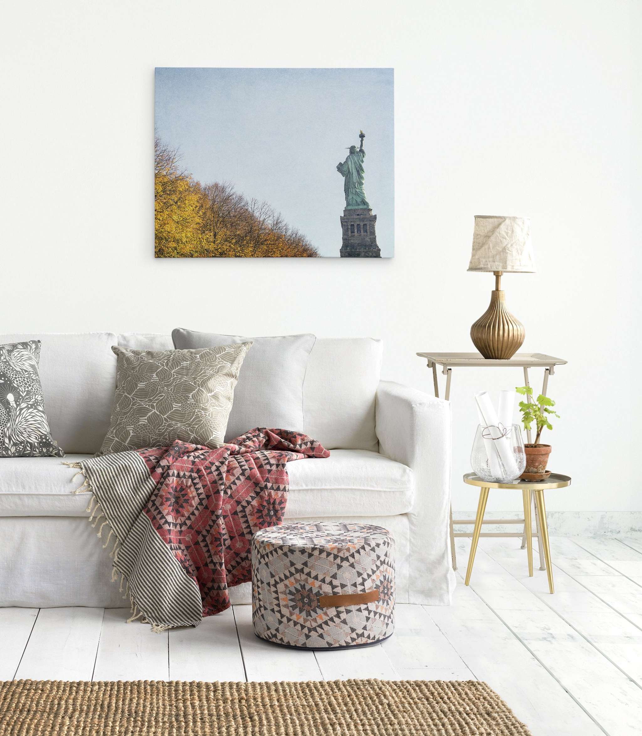 A bright living room features a white couch with patterned cushions and a colorful throw blanket, an ottoman, a side table with a lamp, and a small plant. A wall-mounted picture of the Statue of Liberty on premium artist-grade canvas is visible above the couch. The New York City Canvas Wall Art, 'Liberty in Fall' by Offley Green adds a stylish touch to the décor. Light wood flooring completes the setting.