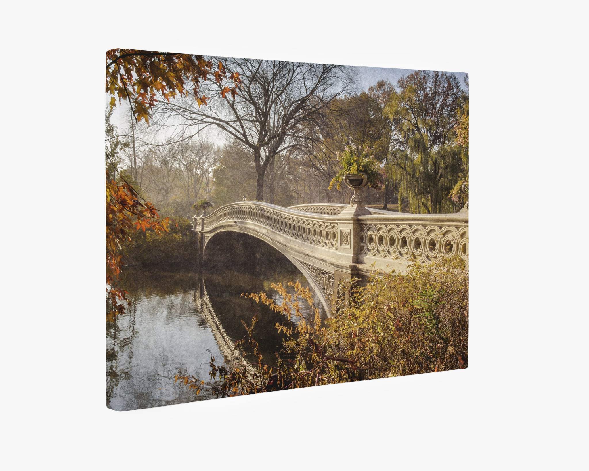 A view of the Bow Bridge in Central Park during autumn showcases the ornate, arched structure adorned with planters, spanning over a calm body of water. Surrounded by colorful fall foliage in shades of orange, yellow, and red with bare trees in the background, it’s perfect for an Offley Green New York City Canvas Wall Art, 'Fall Bow Bridge.'