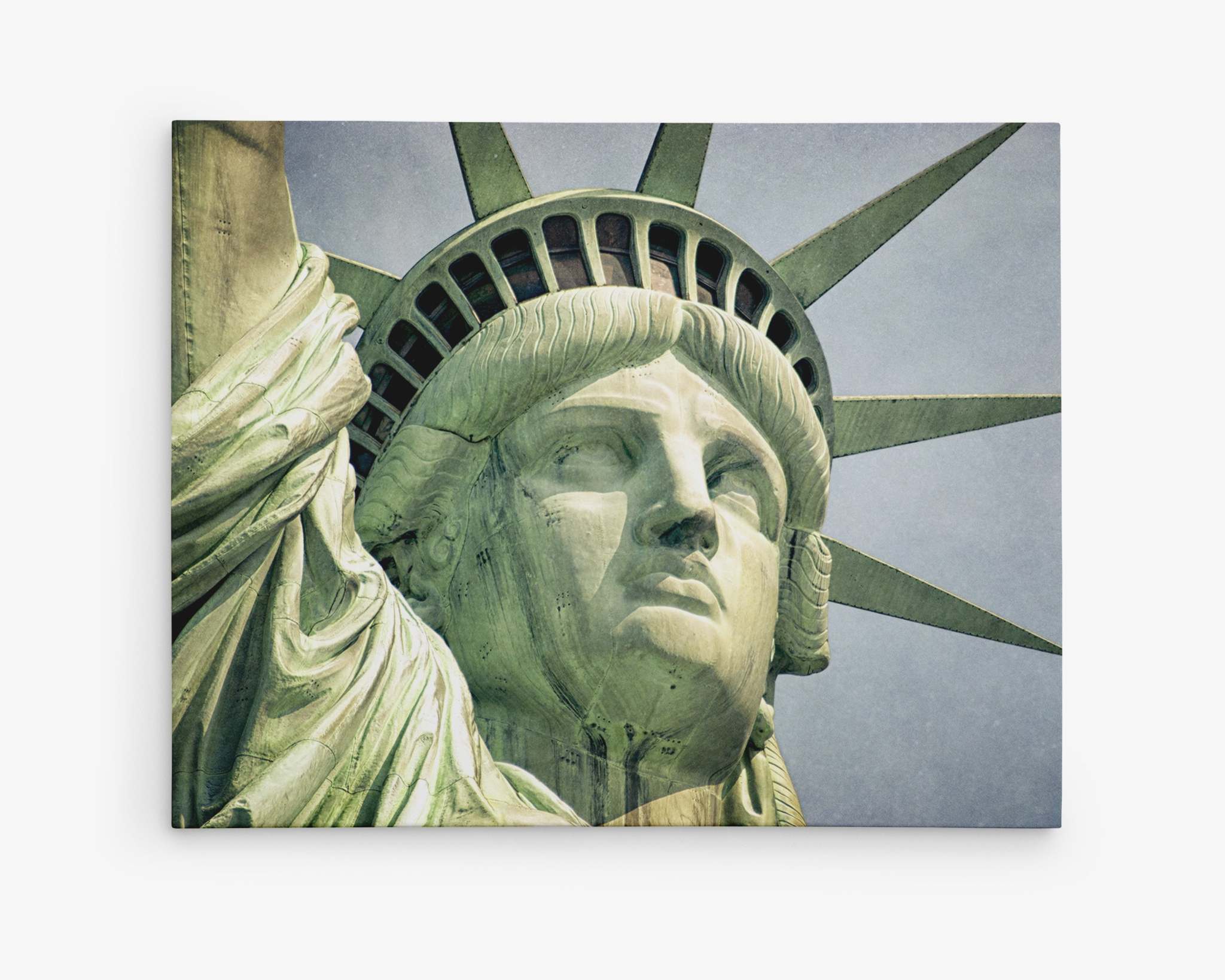 Close-up image of the Statue of Liberty's face and crown, printed on premium artist-grade canvas. The photograph captures the statue's stern expression and the seven rays of the crown against a pale blue sky. The green oxidized copper surface shows detailed texture and historical weathering. This is New York City Canvas Wall Art, 'Face of Liberty' by Offley Green.