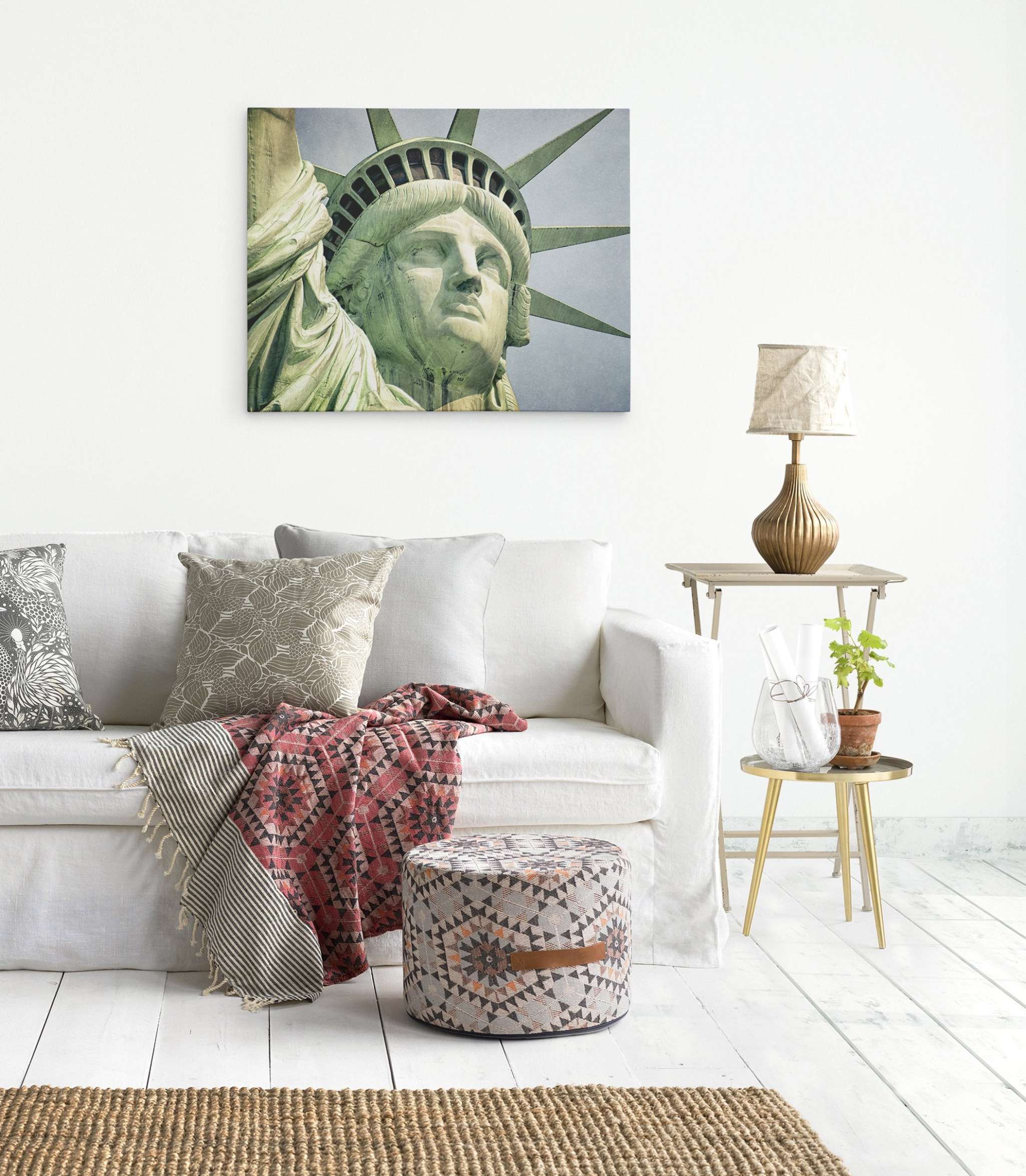 A bright, modern living room with a white sofa adorned with patterned cushions and a colorful throw blanket. Above the sofa, a framed picture of Offley Green's New York City Canvas Wall Art, 'Face of Liberty' on premium artist-grade canvas adds a sophisticated touch. A side table holds a lamp, small plant, and an open book. A patterned ottoman sits on the floor.