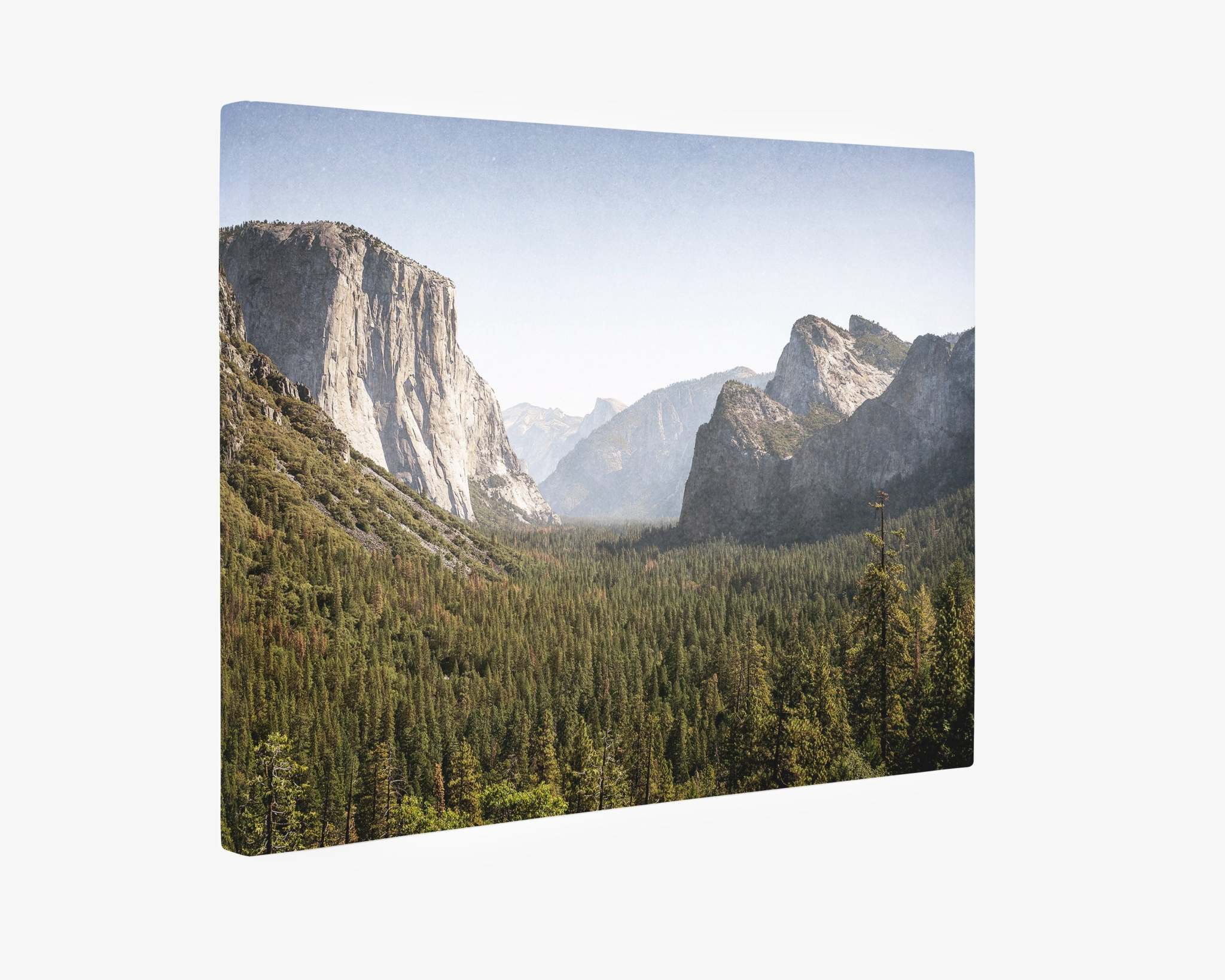 The Northern California Canvas Wall Art, 'Yosemite Valley' by Offley Green showcases a breathtaking view of Yosemite Valley. Towering granite cliffs, including El Capitan on the left, rise above lush green pine forests, with a hazy blue sky overhead, capturing the majestic natural beauty of this iconic part of Yosemite National Park.