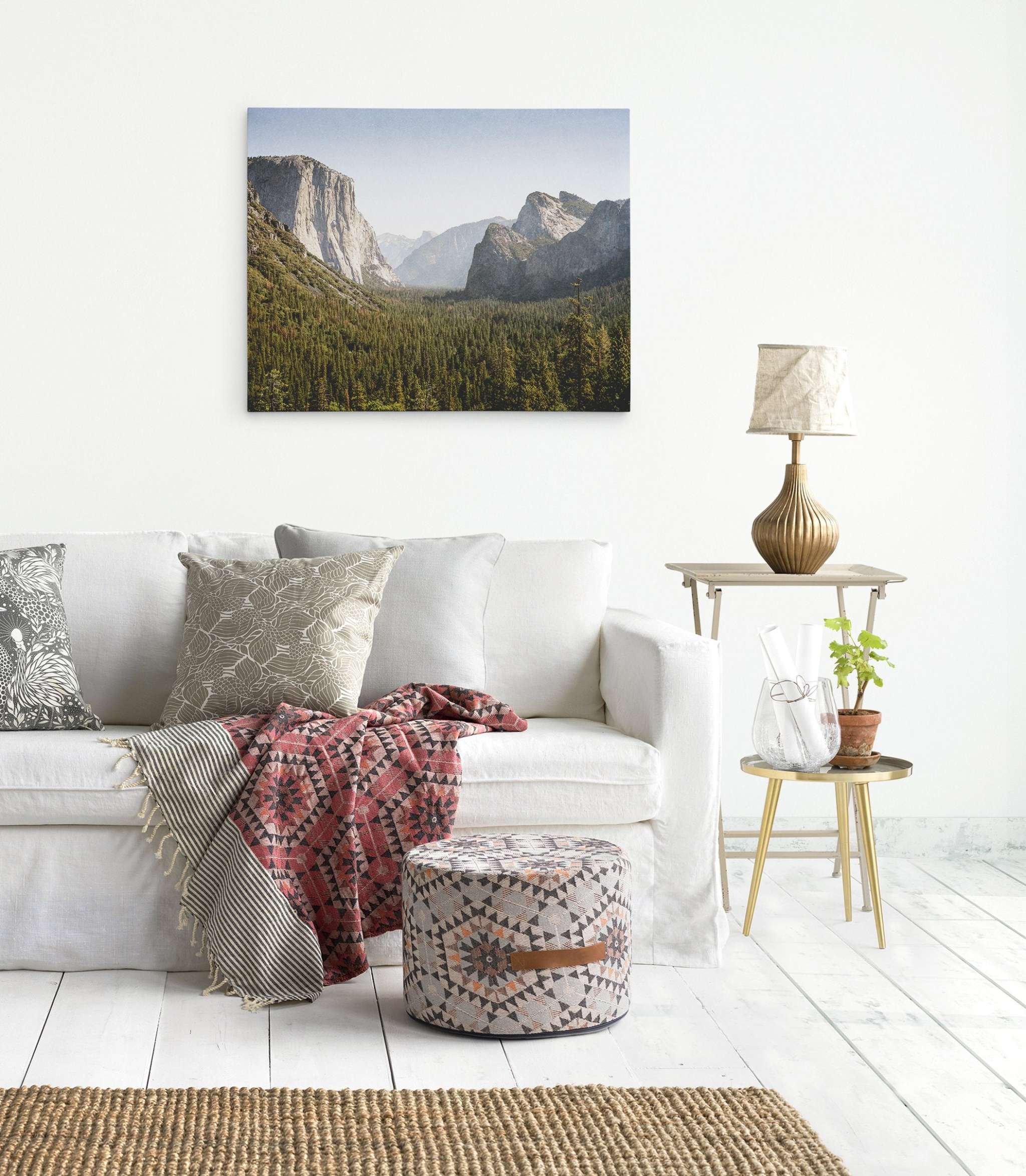 A modern living room features a white sofa with assorted patterned cushions and a red patterned throw blanket. A small table with a gold lamp and plant is adjacent. A round patterned pouf sits nearby, and Offley Green&#39;s &#39;Yosemite Valley&#39; Northern California Canvas Wall Art printed on premium artist-grade canvas hangs on the wall.