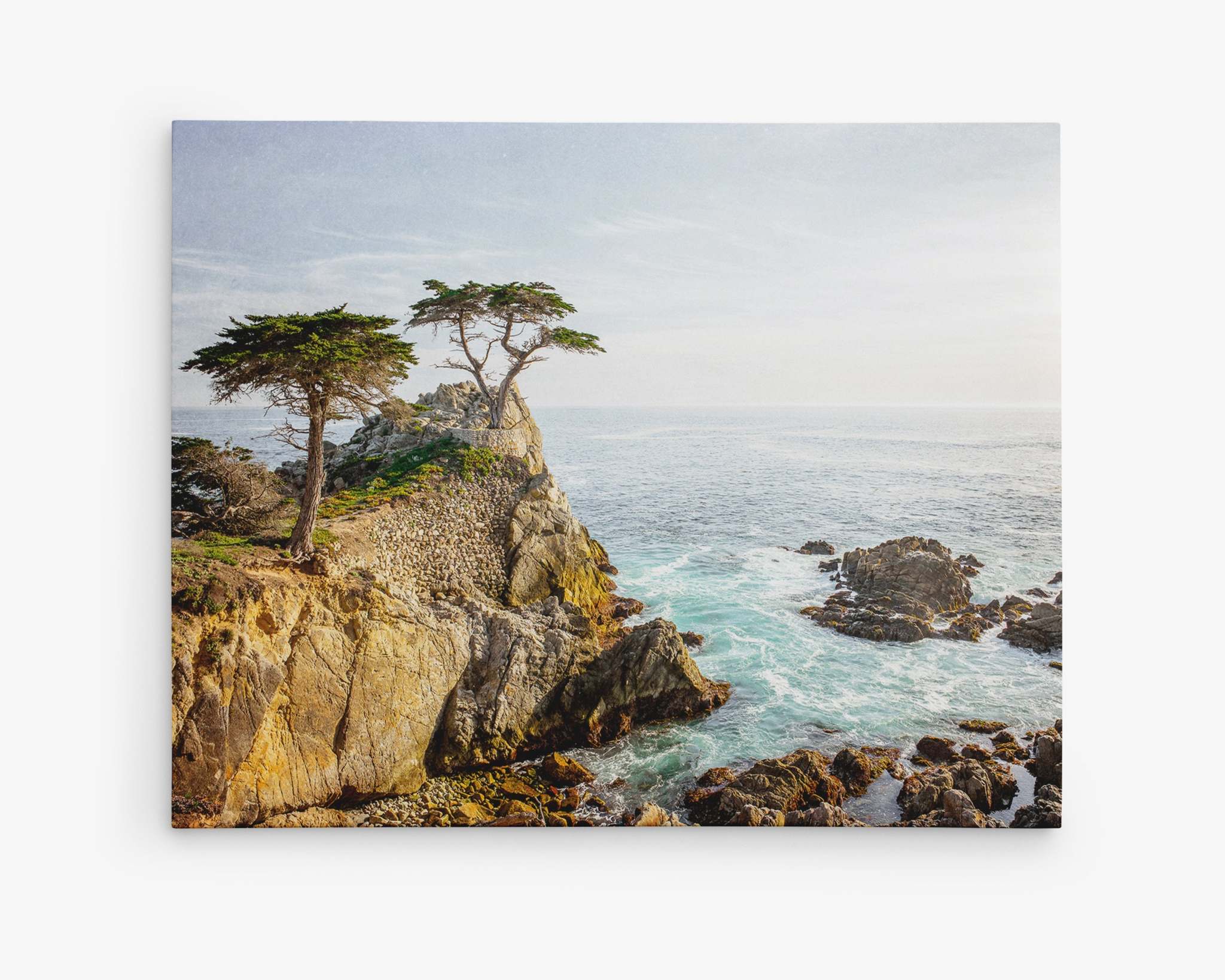 A scenic view of a rocky coastline with a few windswept trees on top of a cliff overlooking the ocean. The sky is clear with a few wispy clouds, and waves crash gently against the rocks below. This tranquil and picturesque setting is perfect for Offley Green's California Coastal Canvas Wall Art, 'Lone Cypress', on premium artist-grade canvas.