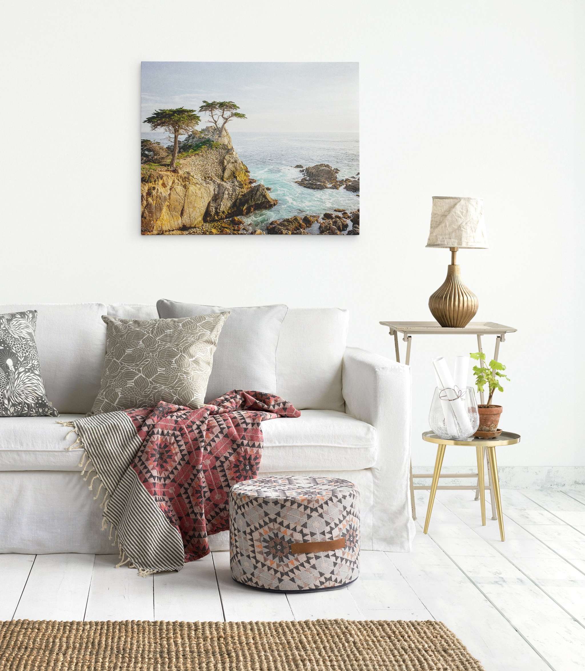 A cozy living room features a white sofa adorned with textured throw pillows and a patterned blanket. A decorative pouf and a side table with a lamp and plant add charm. A landscape painting of California Coastal Canvas Wall Art, &#39;Lone Cypress&#39; by Offley Green hangs on the white wall, completing the serene look.