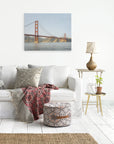 A cozy living room features a white sofa adorned with patterned throw pillows and a colorful blanket. Nearby is a round ottoman with geometric patterns, and a small side table holding a potted plant, glass jug, and table lamp. An Offley Green San Francisco Canvas Wall Art, 'Golden Gate' hangs above.