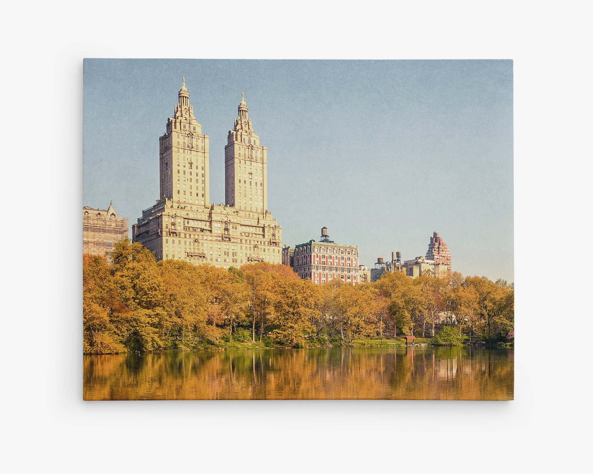 A view of two tall, adjacent buildings with spires, set against a clear blue sky. The buildings are flanked by shorter structures, and there are trees with autumn foliage in the foreground, reflected in a calm body of water below. This scene is captured on the Offley Green New York City Canvas Wall Art, 'Central Park Fall,' offering a ready-to-hang solution for your home.