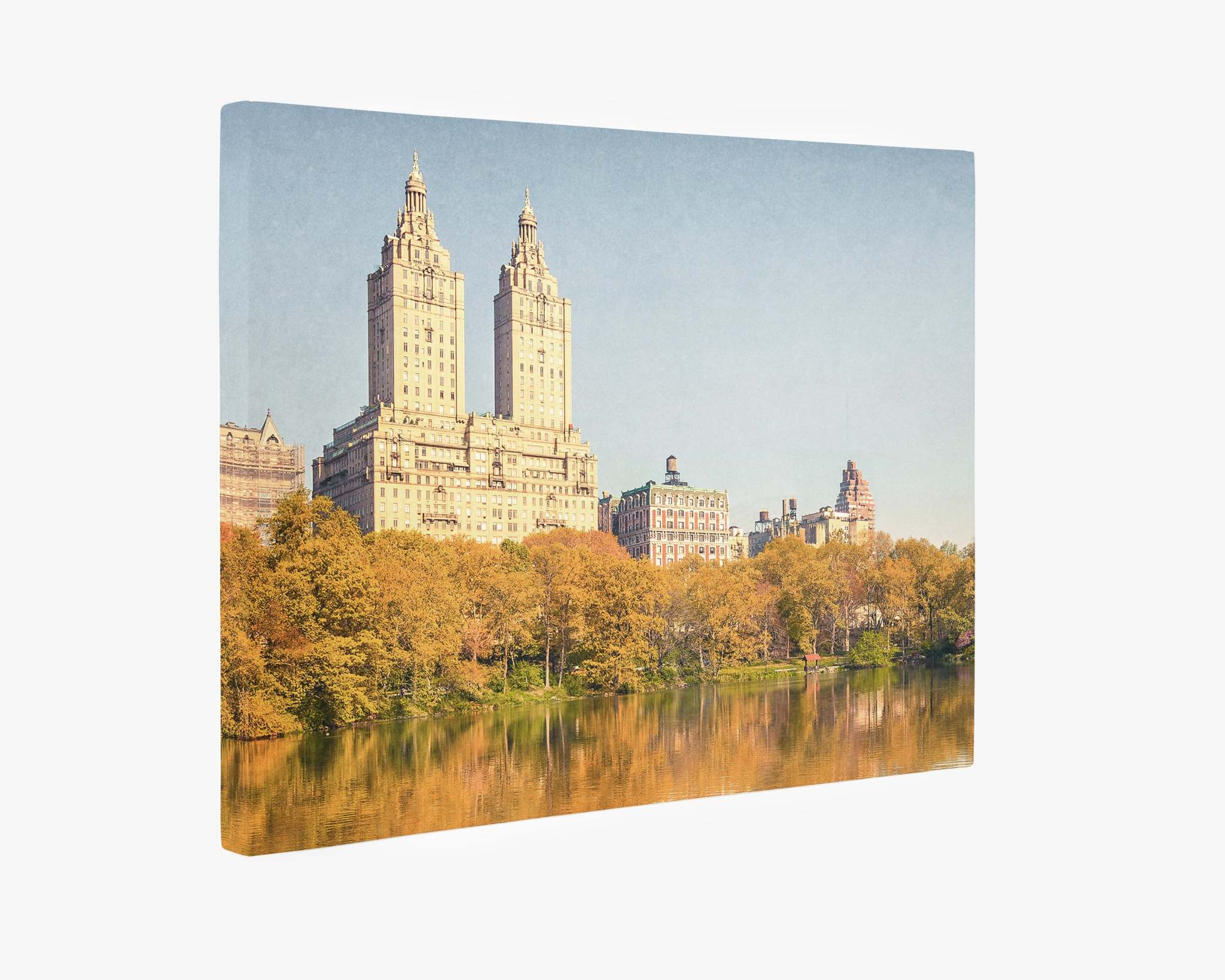 This New York City Canvas Wall Art, 'Central Park Fall' by Offley Green depicts a serene city park scene during autumn. The foreground features a calm lake surrounded by trees with golden foliage, while tall, historic skyscrapers rise against a clear blue sky in the background. This ready-to-hang solution offers premium artist-grade quality for your space.