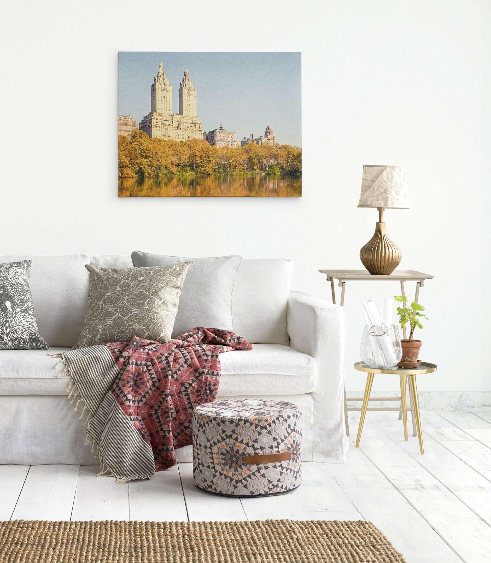 A cozy living room with white walls and floors. There's a white sofa adorned with various patterned pillows and a red geometric throw. A side table holds a lamp and a small plant. Above the sofa, an Offley Green New York City Canvas Wall Art, 'Central Park Fall' serves as the perfect ready-to-hang solution. A patterned pouf is in front.