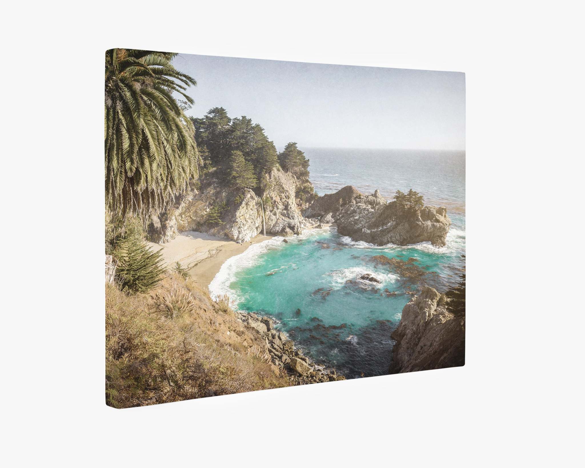 An Offley Green Big Sur Canvas Wall Art, 'Julia Pffeifer,' featuring a secluded beach surrounded by rugged cliffs and lush vegetation. Turquoise waves gently wash onto the shore under a peaceful, clear sky. Dense trees, including a tall palm, cover the cliffside, capturing the essence of Big Sur wall art.