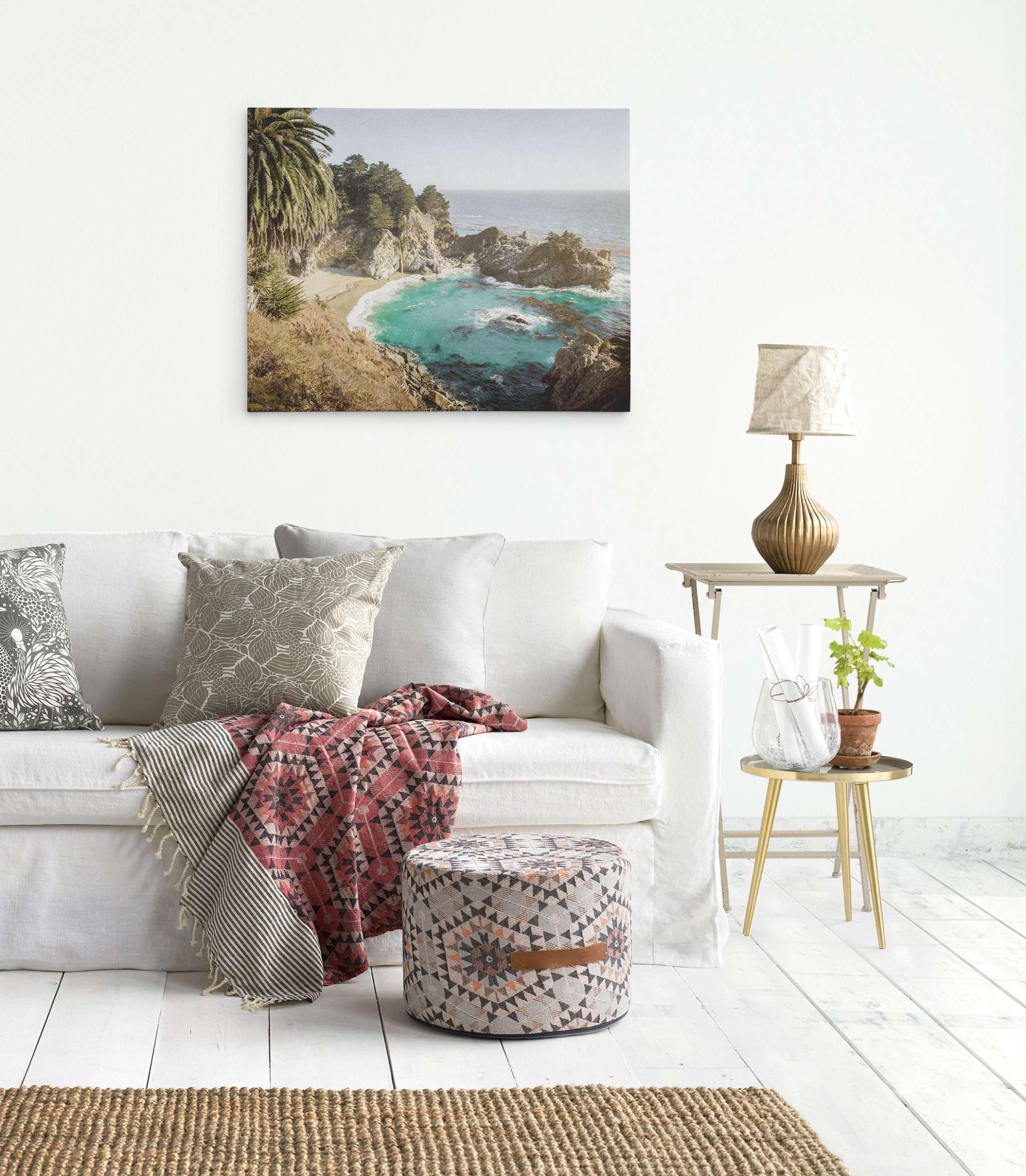 A stylish living room with a white sofa adorned with patterned cushions and a red patterned throw. A round patterned ottoman sits on a textured rug. A side table with a bottle vase, plant, and lamp is nearby. Offley Green&#39;s Big Sur Canvas Wall Art, &#39;Julia Pffeifer&#39; featuring the Pacific Coast Highway waterfall hangs above as a stunning focal point.